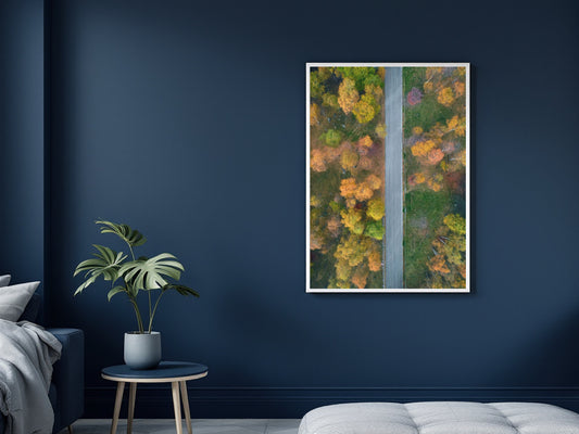 A vertical framed art print depicting an aerial view of a road splitting through vibrant autumn forest foliage, displayed on a dark navy blue wall in a modern bedroom setting.