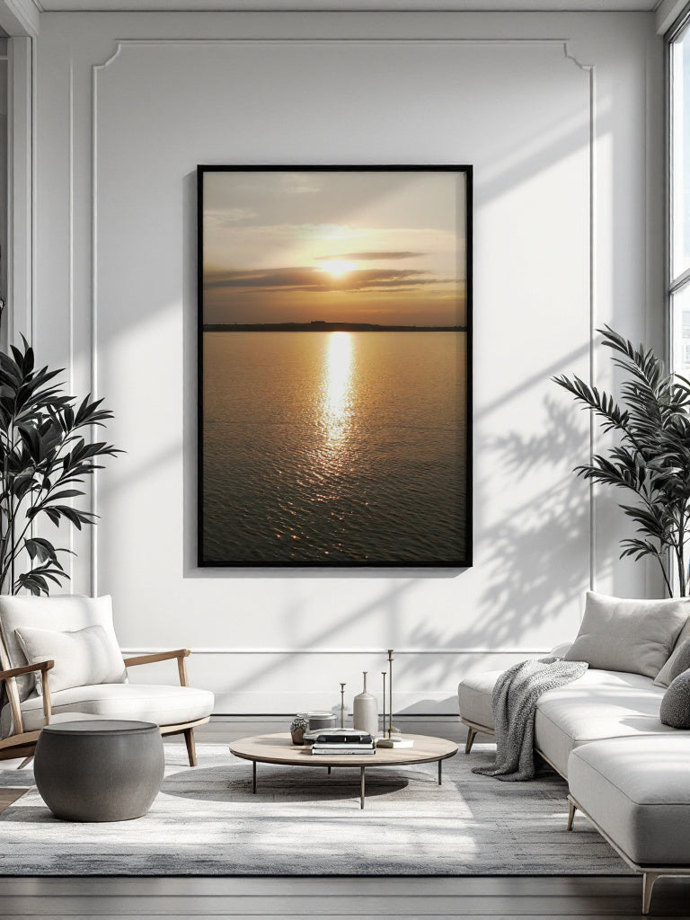 A framed vertical canvas print of a golden sunset over water, displayed on a white paneled wall in a modern, sunlit living room with a sofa, armchair, and coffee table.