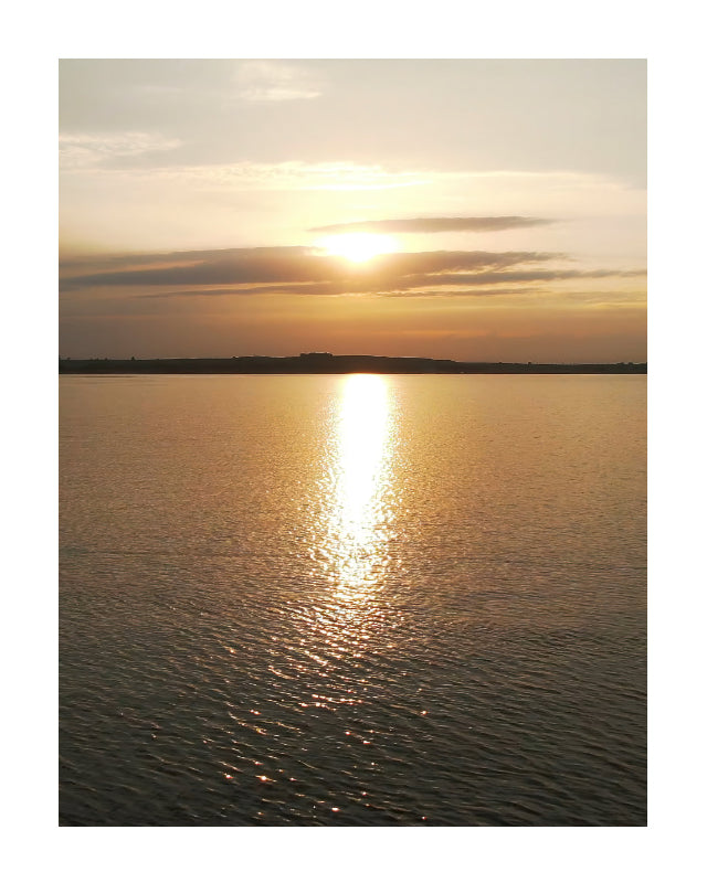 A scenic sunset over a calm body of water, with the sun low on the horizon, golden light reflecting across the surface, and a dark shoreline silhouette in the distance.