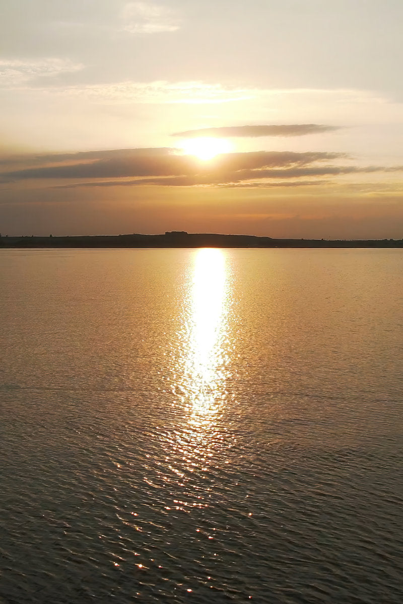 Sun setting over a calm body of water, with golden light reflecting across the surface and a dark shoreline silhouette in the distance.
