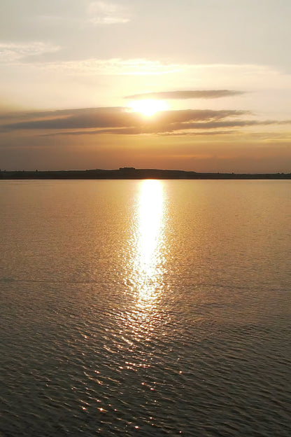 Sun setting over a calm body of water, with golden light reflecting across the surface and a dark shoreline silhouette in the distance.