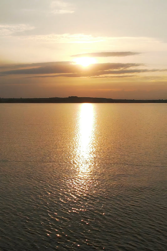 Sun setting over a calm body of water, with golden light reflecting across the surface and a dark shoreline silhouette in the distance.