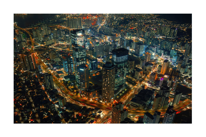 Aerial night photograph of a dense city skyline with illuminated skyscrapers, glowing streetlights, and a dark urban background, presented with a white border.