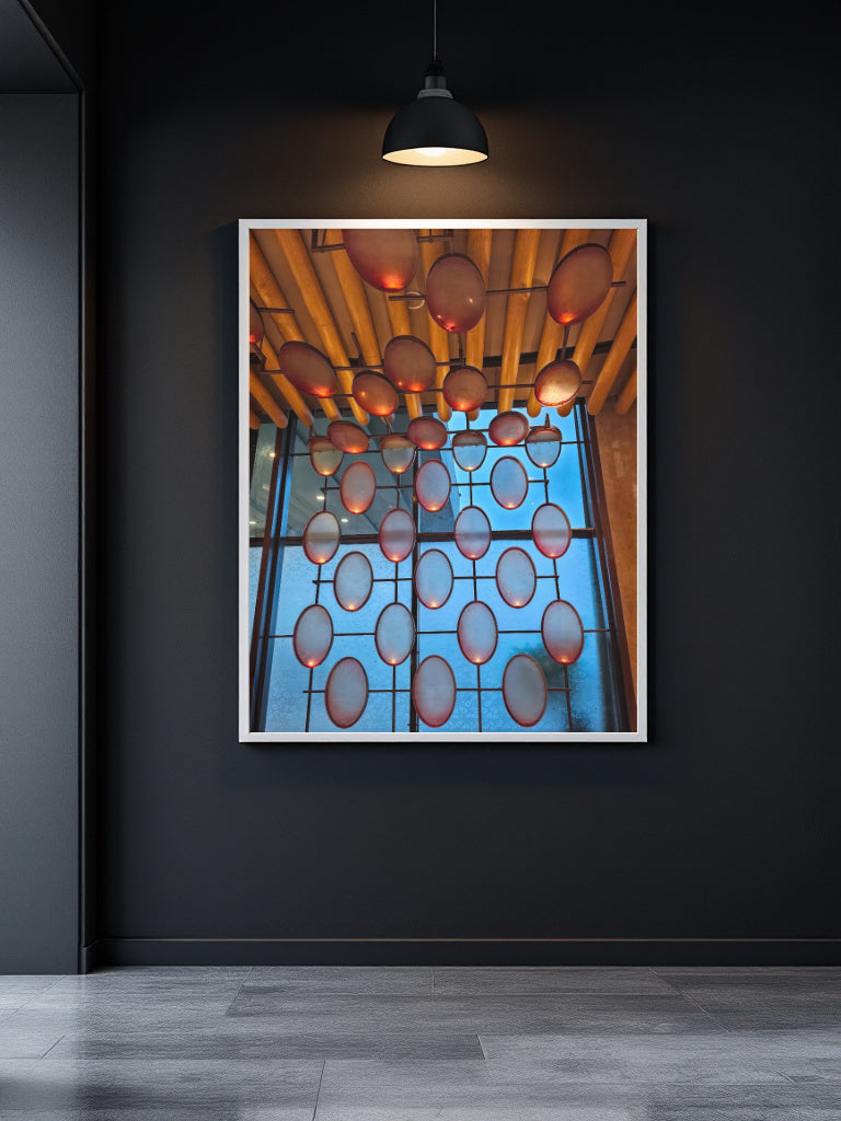 A framed abstract art print featuring a grid of glowing orange and red circular shapes against a bright blue background, hanging on a dark gray wall in a modern room with a black pendant light above.
