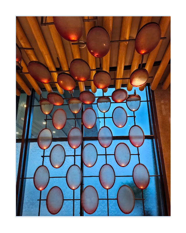 A modern architectural interior view looking upward at a grid of oval glass panels glowing with warm amber light, suspended beneath a slatted wooden ceiling.
