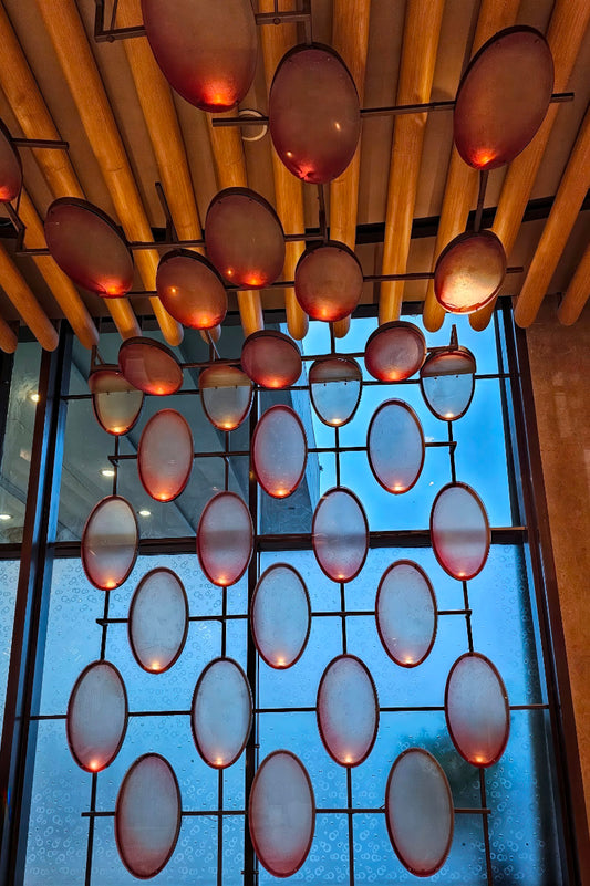 A decorative architectural glass screen or wall panel with multiple oval frosted glass panels set in a dark metal grid, backlit by warm light from hanging oval lamps beneath a slatted wood ceiling.