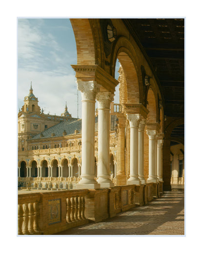A scenic architectural photograph of the Plaza de España in Seville, Spain, featuring a grand colonnade with white columns and arches framing a courtyard and historic building under a blue sky.