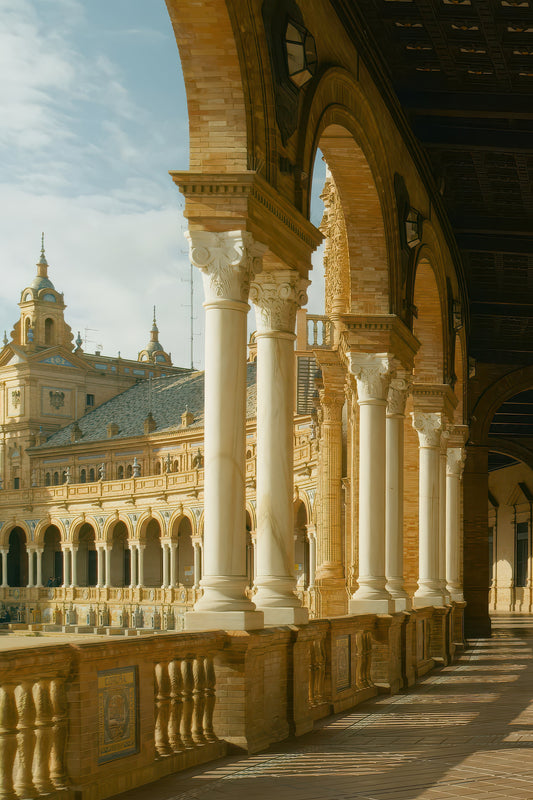 A scenic architectural photograph of the Plaza de España in Seville, Spain, featuring a warm golden-beige brick colonnade with white columns overlooking a grand courtyard under a blue sky.