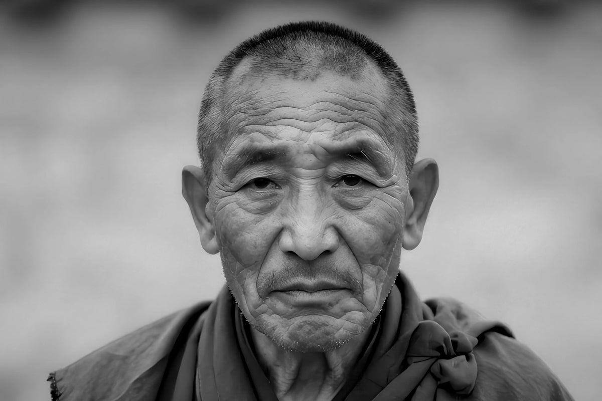 A black and white close-up portrait of an elderly man with a short hair, wearing a dark draped garment, set against a soft blurred gray background.
