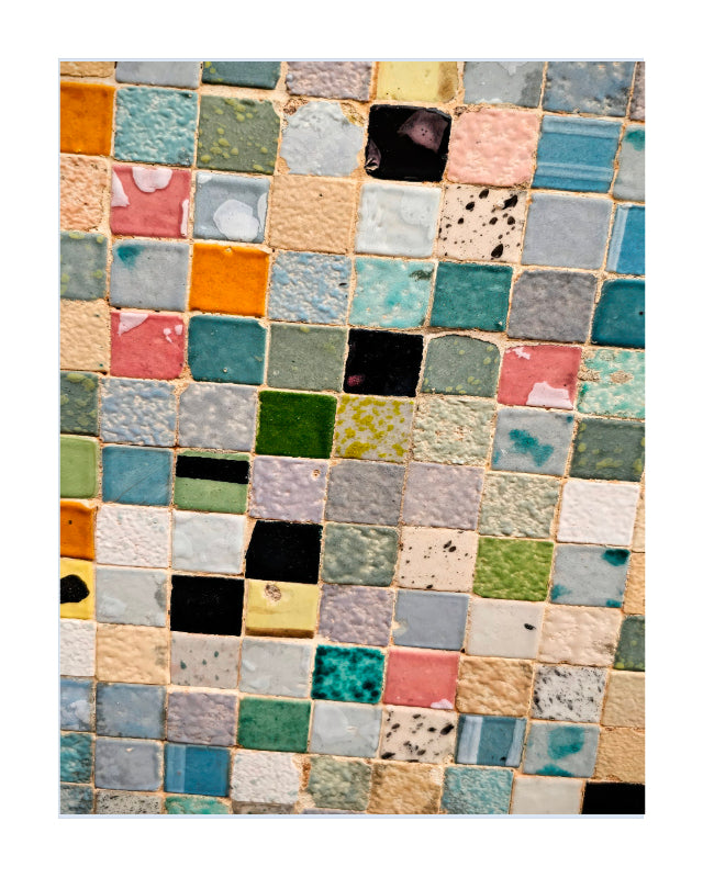 A framed abstract patchwork art print with a colorful grid of square tiles hangs on a white wall above a wooden desk with plants, a white chair, and a black lamp.
