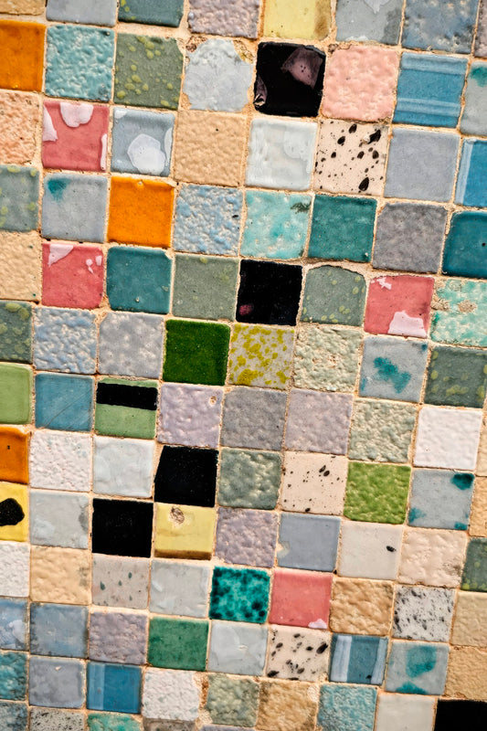 A close-up of a mosaic tile pattern made of many small square tiles in assorted pastel and earthy colors including blue, green, pink, beige, yellow, black, and white.