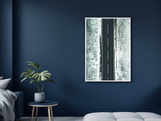 A vertical framed wall art print showing a narrow road running through a snowy forest, displayed on a dark navy blue wall in a modern bedroom setting next to a plant and bed.