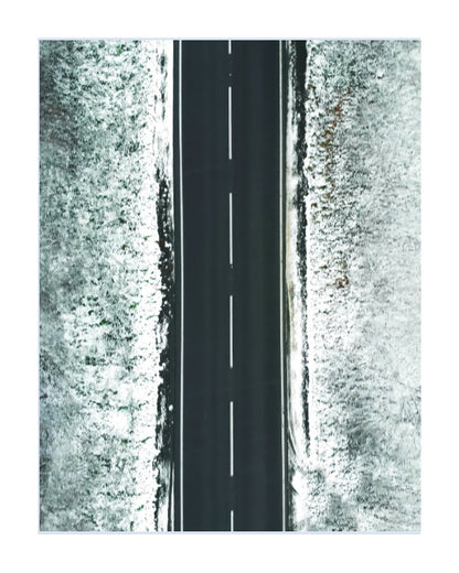 A minimalist aerial photograph of a straight black road with a dashed white centerline, flanked by textured white snowy fields on both sides, presented on a white background.