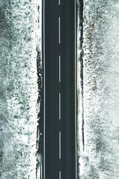Aerial view of a straight paved road with a dashed centerline, flanked by snow-covered ground on both sides.