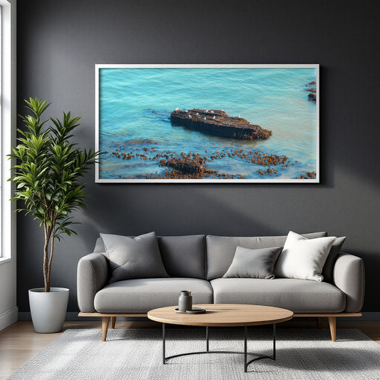 A framed wall art print depicting a shipwreck in turquoise ocean water, displayed above a gray sofa in a modern living room setting.
