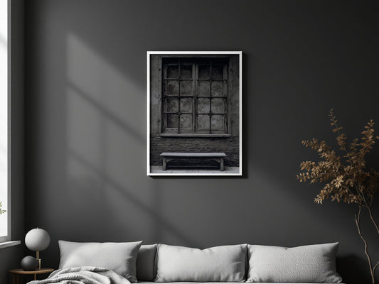 A framed black and white art print of an old window and bench, displayed on a dark gray wall above a modern gray sofa in a styled living room.