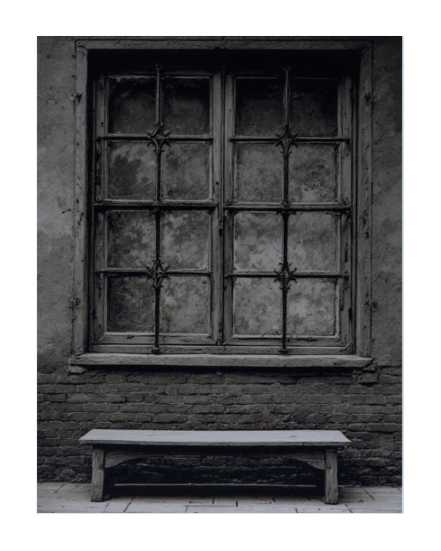 Black and white photograph of an old wooden window with iron bars set in a brick wall, with a simple wooden bench below it.