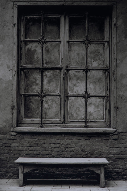 A dark, moody photograph of an old, weathered wooden window with multiple panes and a rustic brick wall behind it, with a simple wooden bench in the foreground.
