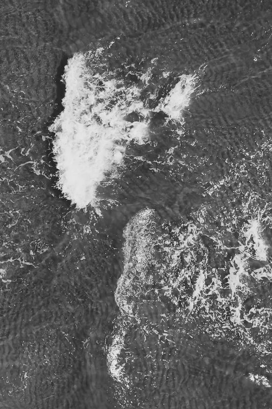 Black and white aerial photograph of ocean waves breaking, showing textured dark water and bright white foam.