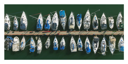Aerial view of multiple white sailboats with blue accents docked in rows along a wooden pier over dark green water.