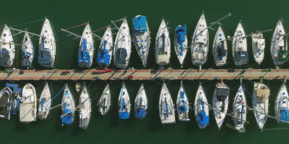Aerial view of a marina with many sailboats docked side by side in green water along a wooden pier.