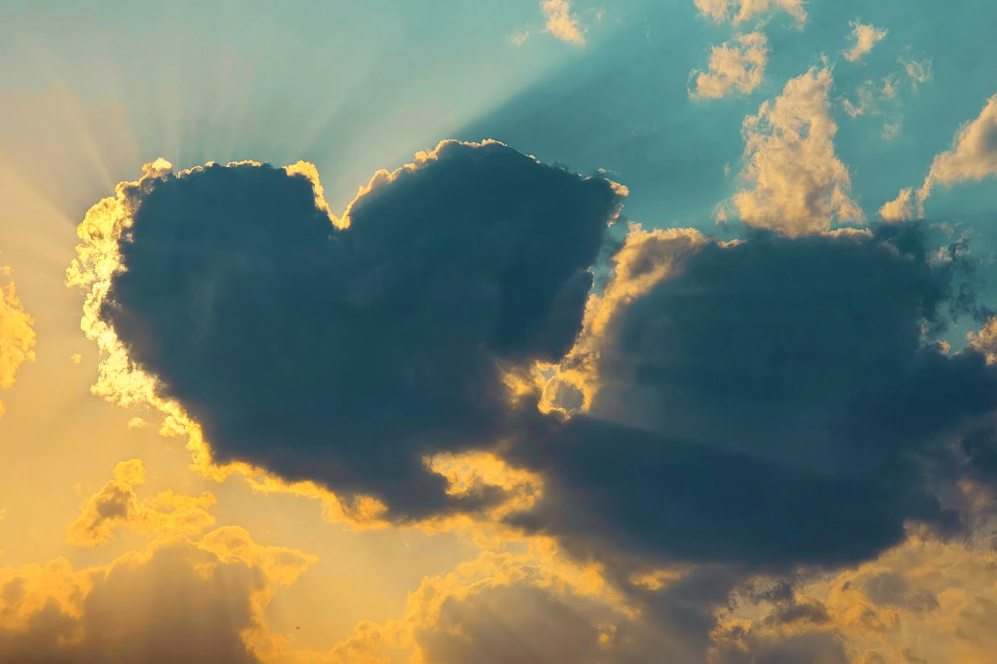 A dramatic sky scene with a heart-shaped cloud silhouette backlit by golden sun rays, surrounded by blue sky and warm yellow-orange clouds.