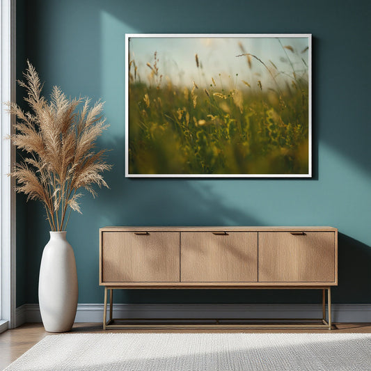 A framed art print depicting a soft-focus golden grassy field hangs on a teal wall above a light wood sideboard, with a tall white vase of pampas grass to the left.