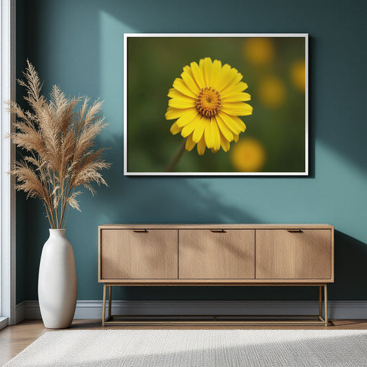 A framed square wall art print showing a bright yellow flower on a blurred green background, displayed above a light wood console table in a styled interior with a teal wall, vase of pampas grass, and rug.