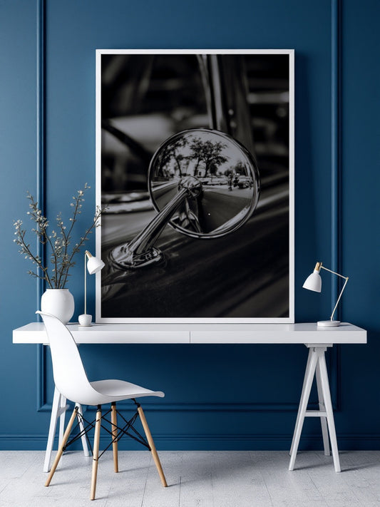 A large black and white framed art print of a vintage car side mirror hangs on a dark blue paneled wall above a white desk with a chair and lamp.