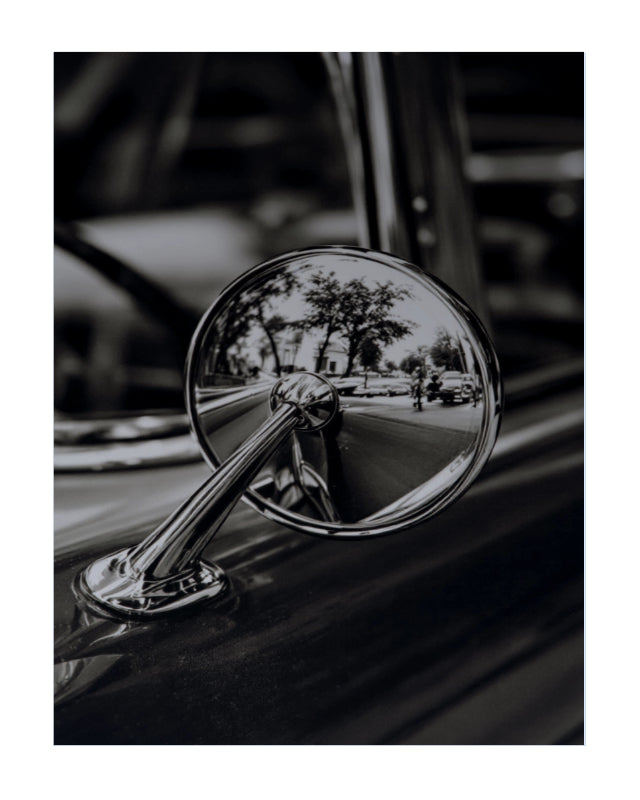 A black and white close-up photograph of a vintage car's chrome side-view mirror reflecting a street scene with trees and a car.