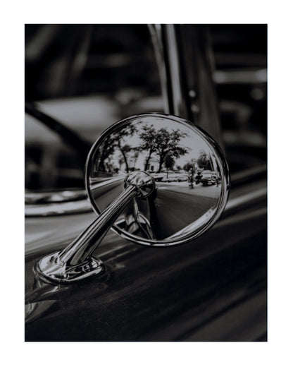 A black and white close-up photograph of a vintage car's chrome side-view mirror reflecting a street scene with trees and a car.