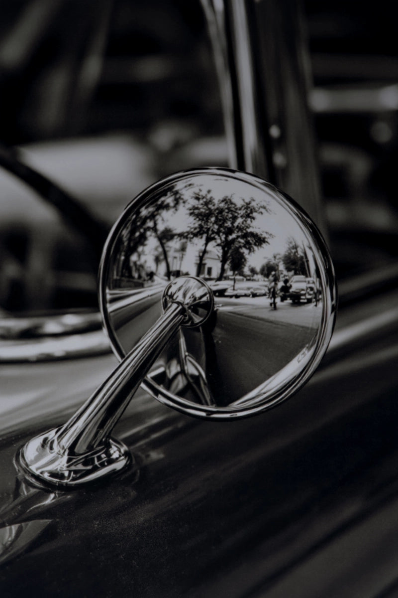A close-up black and white photograph of a classic car's chrome side-view mirror attached to the fender, reflecting a blurred street scene.