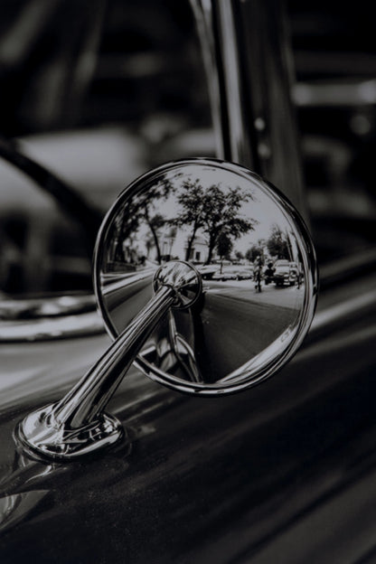 A close-up black and white photograph of a classic car's chrome side-view mirror attached to the fender, reflecting a blurred street scene.