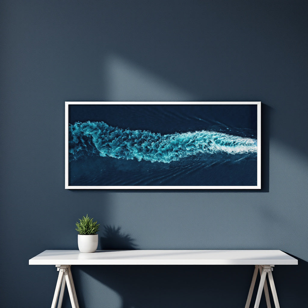 A framed panoramic art print of a turquoise ocean wave on a dark navy wall above a white console table with a small potted plant.