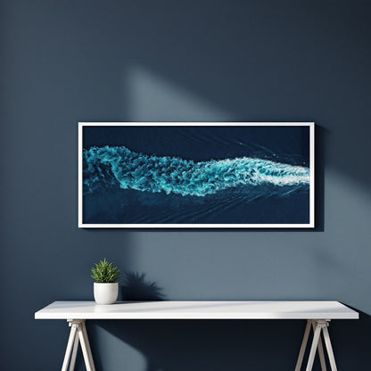 A framed panoramic art print of a turquoise ocean wave on a dark navy wall above a white console table with a small potted plant.