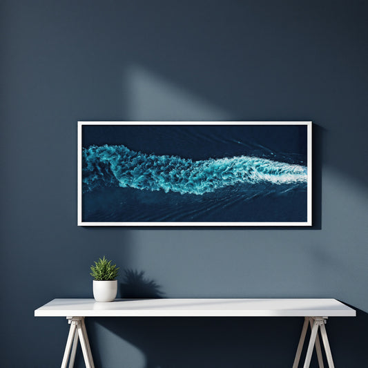 A framed panoramic art print of a turquoise ocean wave on a dark navy wall above a white console table with a small potted plant.