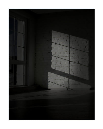 A moody black and white photograph of a room corner with a window casting dramatic light and shadow across a white brick wall and dark floor.