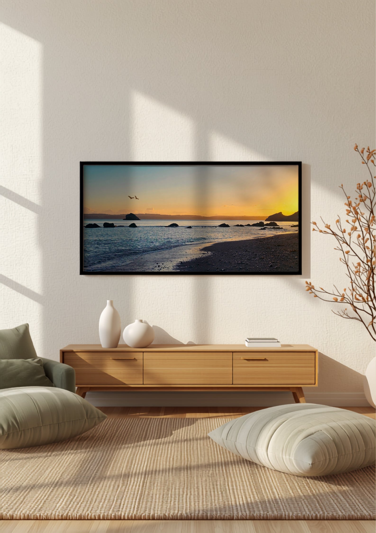 A framed wall art print showing a beach sunset scene, displayed in a modern living room setting above a light wood console table with decorative vases and cushions.