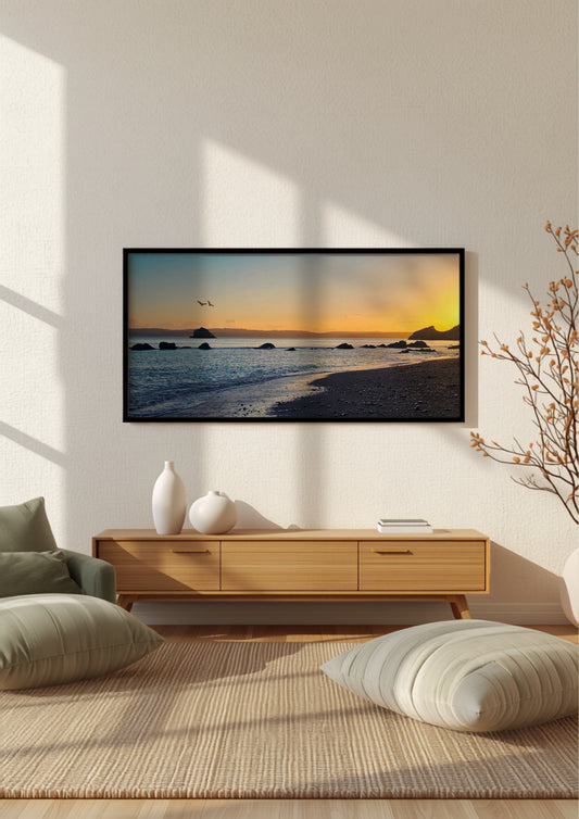 A framed wall art print showing a beach sunset scene, displayed in a modern living room setting above a light wood console table with decorative vases and cushions.
