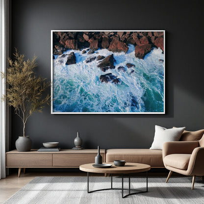 A framed square wall art print showing an aerial view of turquoise ocean waves crashing around dark rocky shores, displayed above a beige sofa in a modern living room setting.