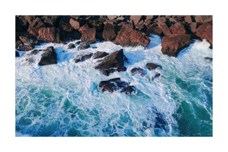 A square photographic print of an aerial coastal scene showing turquoise ocean water with white surf crashing around dark reddish-brown rocks, centered on a white background.