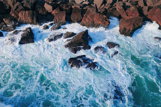 Aerial view of turquoise ocean waves crashing against dark reddish-brown rocks, with white sea foam surrounding the stones.