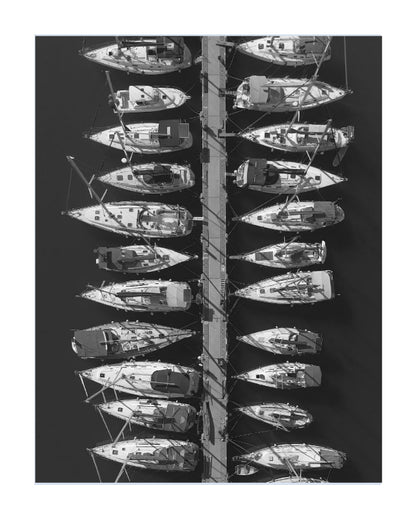 Black and white aerial photograph of multiple sailboats docked side-by-side along a narrow channel, presented as a square art print with a white border on a white background.