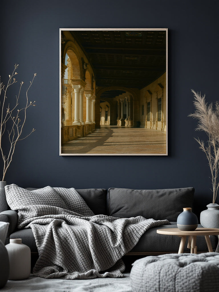 A framed art print depicting a warm-toned classical arcade corridor hangs on a dark navy wall above a gray sofa in a styled living room setting.