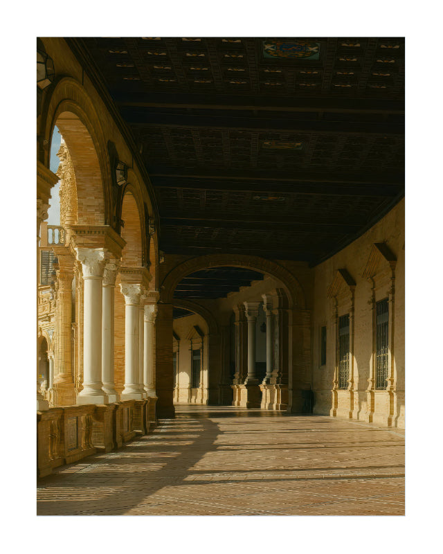 A scenic photograph of a grand architectural colonnade with arches, columns, and a dark coffered ceiling, casting long shadows across a stone floor.