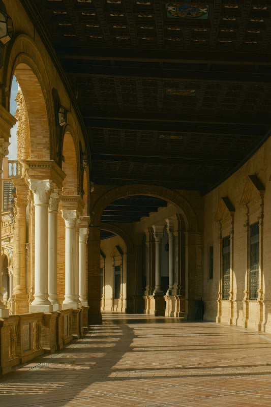 A long covered arcade corridor with repeating arches and columns, lit by warm sunlight casting long shadows across a tiled floor. The architecture features tan stone walls and a dark coffered ceiling.