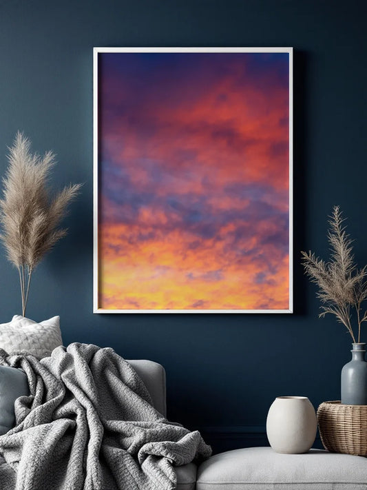 A framed vertical art print depicting a vibrant sunset sky with orange, yellow, and purple clouds, displayed on a dark navy wall above a modern gray sofa with a textured throw blanket and decorative dried grasses.