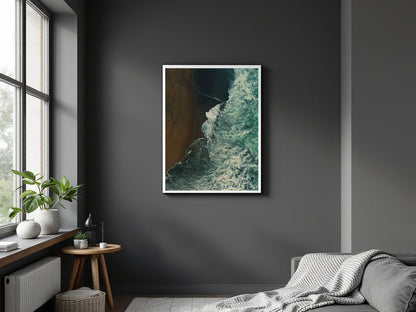 A framed vertical art print depicting a dramatic coastline with dark teal water and a brown sandy shore, displayed on a dark gray wall in a modern bedroom setting.
