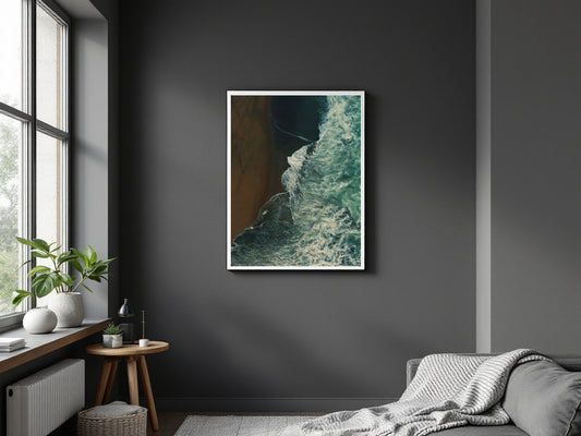 A framed vertical art print depicting a dramatic coastline with dark teal water and a brown sandy shore, displayed on a dark gray wall in a modern bedroom setting.