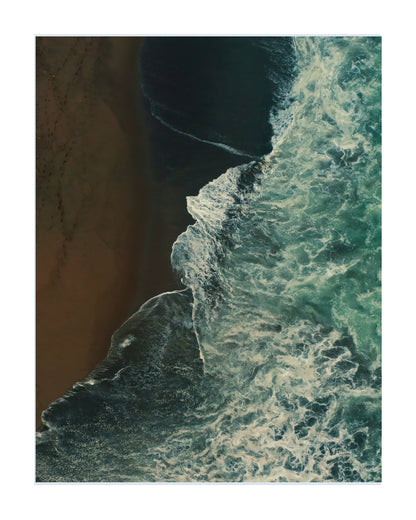An aerial photograph of a coastline showing dark teal ocean waves with white foam crashing against a brown sandy beach, presented as a square art print with a white border.